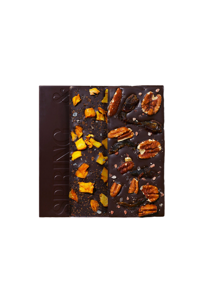 A bar of dark chocolate from the Spice Market Collection by Spring & Mulberry is divided into three sections. The left section is plain, embossed with the word "SPRING," the middle is topped with naturally sweet dried mango pieces, and the right section has a variety of nuts and seeds.