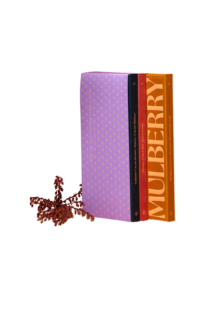 A stack of three chocolates aligned vertically against a neutral background. The chocolates are titled "Spice Market Collection," "Meetings + Conversations," and "MULBERRY" from left to right. Beside the books lies a dried flower arrangement, evoking vibes as naturally sweet as tropical fruits by Spring & Mulberry.