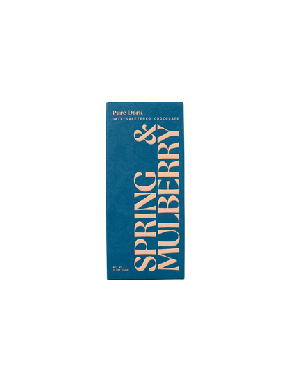 A rectangular chocolate bar package with a dark blue background features "SPRING & MULBERRY" printed in large light pink letters vertically. At the top, it says "Pure Dark Date-Sweetened Chocolate." Crafted with organic cacao, the net weight of 3.2 oz (90 g) is indicated at the bottom left.