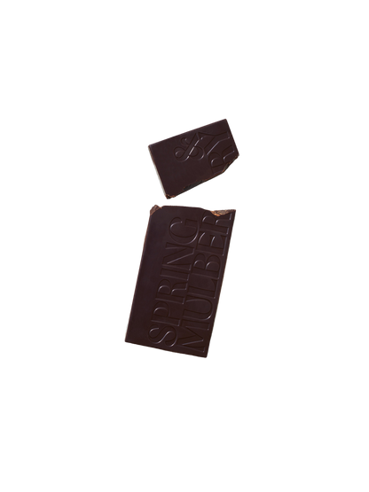 A bar of Pure Dark Date-Sweetened Chocolate with a small broken-off piece on a black background. The larger section of the chocolate bar has the words "Spring & Mulberry" embossed on it, showcasing its rich, date-sweetened chocolate origin.