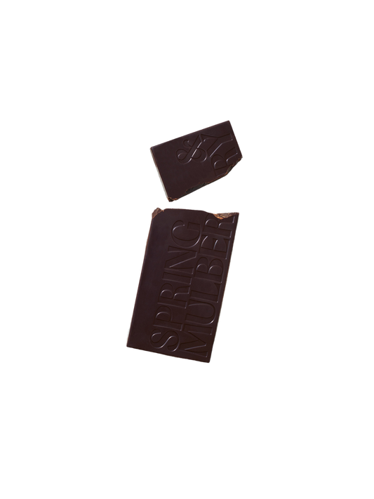 A bar of Pure Dark Date-Sweetened Chocolate with a small broken-off piece on a black background. The larger section of the chocolate bar has the words "Spring & Mulberry" embossed on it, showcasing its rich, date-sweetened chocolate origin.