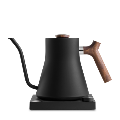 The Fellow Wholesale Stagg EKG Electric Kettle is a sleek, matte black appliance with a minimalist design. It features a precise gooseneck spout, a wooden handle, and a wooden knob on the lid. The kettle offers variable temperature control and sits on a black square base.