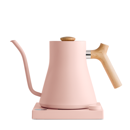 Introducing the Stagg EKG Electric Kettle by Fellow Wholesale: a sleek, modern pour-over kettle featuring a matte pink finish and a wooden handle. This kettle boasts a precisely engineered gooseneck spout and rests on a matching pink base, ensuring perfect pouring every time.