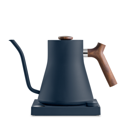 A matte black Stagg EKG Electric Kettle from Fellow Wholesale, featuring a wooden handle and a gooseneck spout, rests on its matching base. The minimalist and modern design seamlessly combines metal and wood elements, offering a sleek, functional appearance along with variable temperature control for perfect brewing precision.