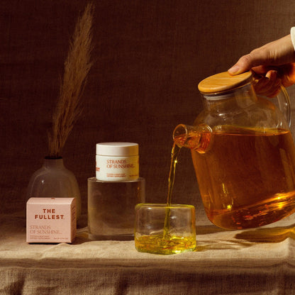 A hand pours tea from a glass teapot into a matching glass cup. Nearby, two jars labeled "Strands of Sunshine" by THE FULLEST, one in pink and the other in white, boast medicinal saffron and offer anxiety support. The jars rest on translucent blocks while a vase with dried grass decorates the textured surface in the background.