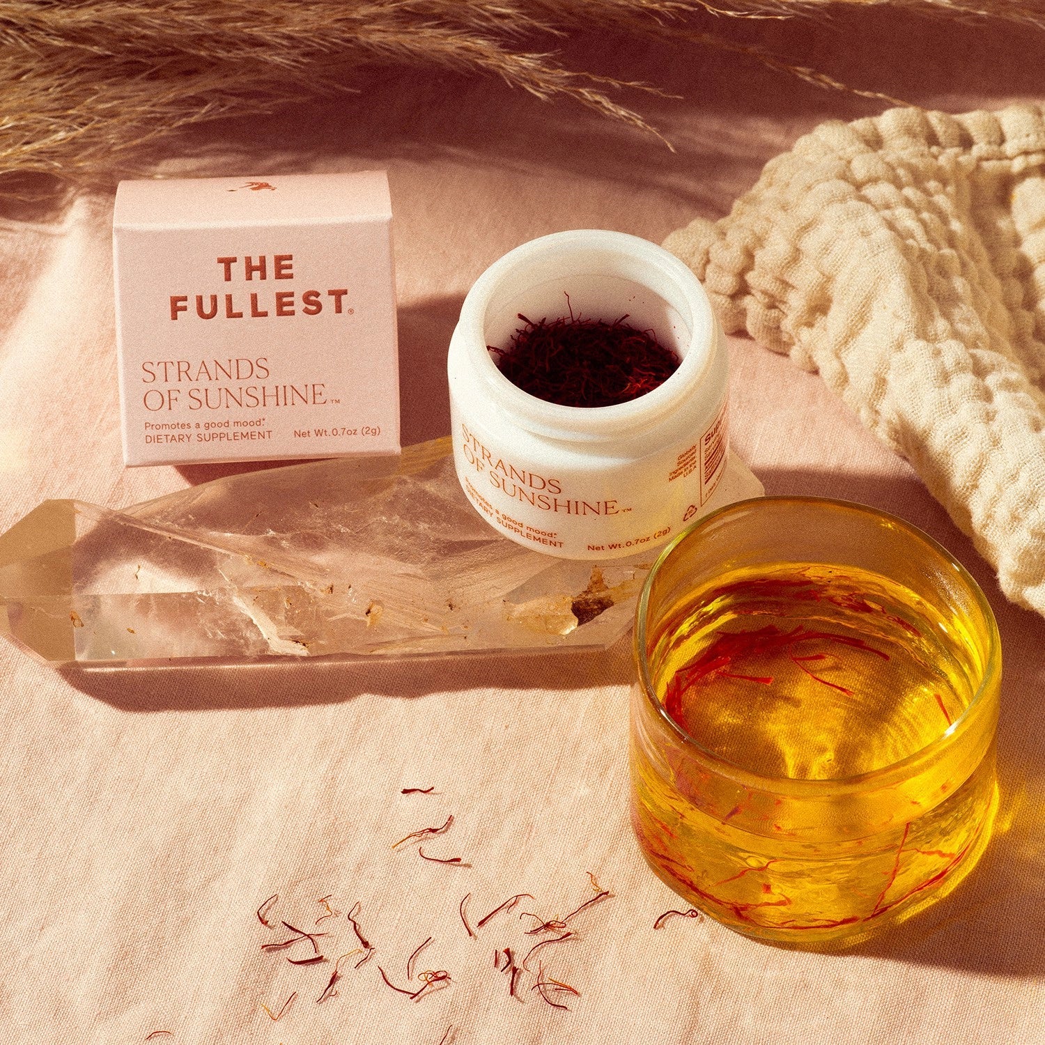 In a pink and beige setting, a variety of wellness products are displayed, including a box from THE FULLEST, an open white jar labeled "Strands of Sunshine" with medicinal saffron threads spilling out, a crystal, and a glass of saffron-infused water. These components collectively provide natural anxiety support through the power of saffron.