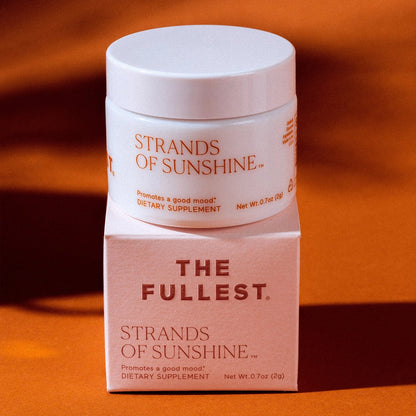 A small jar labeled "Strands of Sunshine" by THE FULLEST is placed on top of a white box. Both the jar and box indicate it is a dietary supplement that "Promotes a good mood." Infused with medicinal saffron, the image features a warm, orange-toned background.