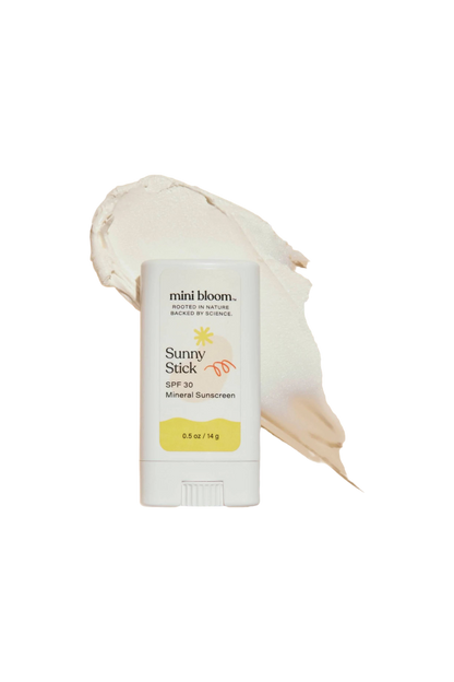 The mini bloom Sunny Stick Mineral Sunscreen SPF 30 is displayed with its cap off against a black background. Behind it, a smear of white sunscreen highlights its texture, showcasing this baby-friendly sunscreen. The stick offers 0.5 oz (14 g) of protective formula.