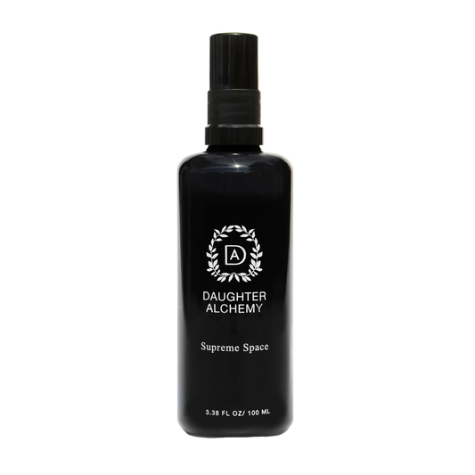 A black spray bottle with a black cap labeled "Daughter Alchemy" and "Supreme Space® Botanical Protective Spray". The bottle also features a circular logo with the letters "DA" in the center. Infused with organic white sage, the volume is listed as 3.38 fl oz / 100 ml.