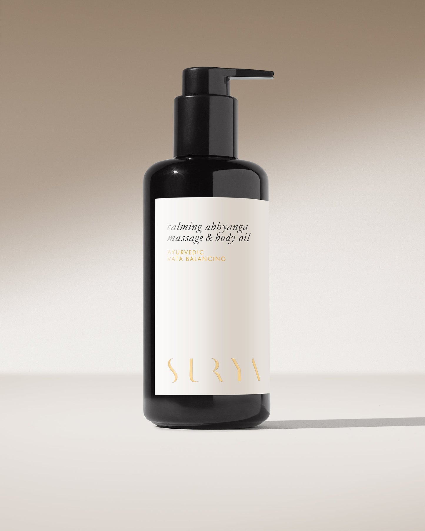 A black pump bottle labeled "Calming Massage & Body Oil" with the brand name "Surya." Infused with Ashwagandha Root and made from organic ingredients, it stands against a gradient background ranging from light to dark beige.