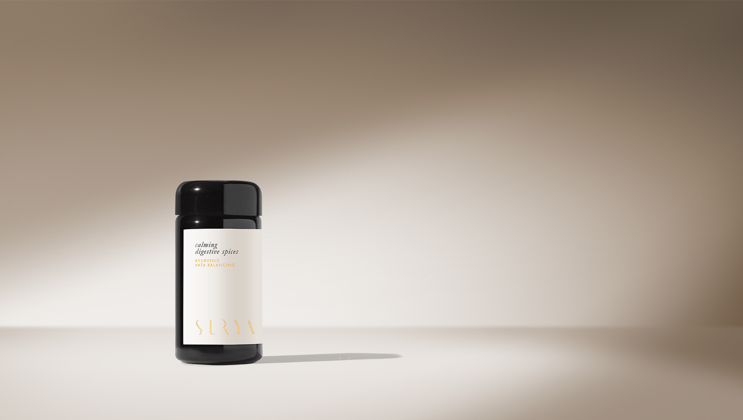 A cylindrical, black-capped bottle labeled "Surya" stands upright against a soft gradient beige background. The bottle, indicated as a Calming Digestive Spice Blend featuring organic ingredients and a minimalistic design, is positioned in a well-lit spotlight from the left.