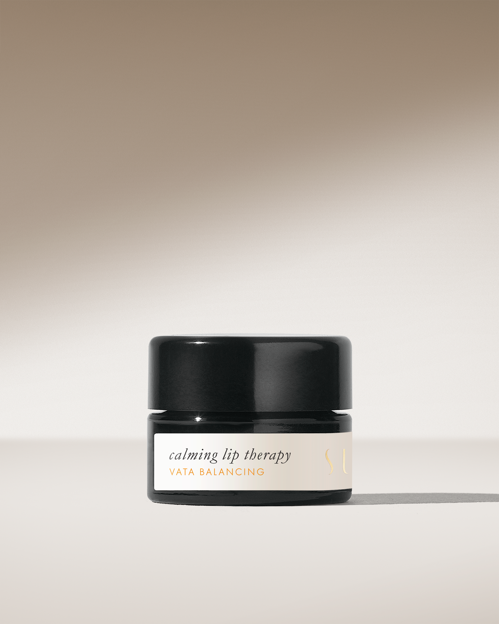 A small black jar labeled "calming lip therapy VATA BALANCING" from the brand Surya sits against a gradient beige background. The jar casts a soft shadow on the surface. The minimalistic and elegant packaging promises a creamy formula made with organic ingredients for effective moisturizing.