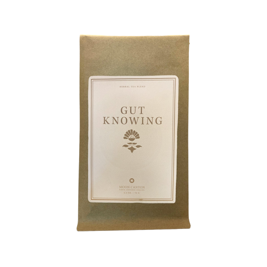 A brown paper package featuring a white label inscribed with "Gut Knowing Tea Blend" and adorned with a floral emblem. The label also presents "HERBAL TEA BLEND" at the top and "Moon Canyon Healing Los Angeles" at the bottom, emphasizing its support for mood and the gut-brain axis. The image is set against a black background.