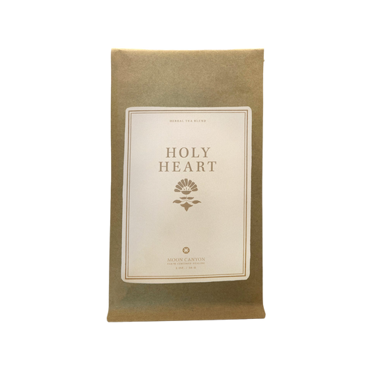 A brown paper pouch with a minimalist and elegant design features a white label at its center. The label prominently displays "Holy Heart Tea Blend" in bold letters, with “Herbal Tea Blend” elegantly printed above it. Below, the small logo reads “Moon Canyon Healing” along with additional text. 