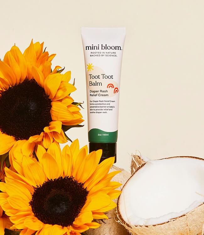 The image features a tube of mini bloom Toot Toot Diaper Rash Balm, a diaper rash balm that creates a protective barrier and soothes irritated skin. The tube is positioned between two sunflowers on the left and a cracked open coconut on the right, all set against a light beige background.