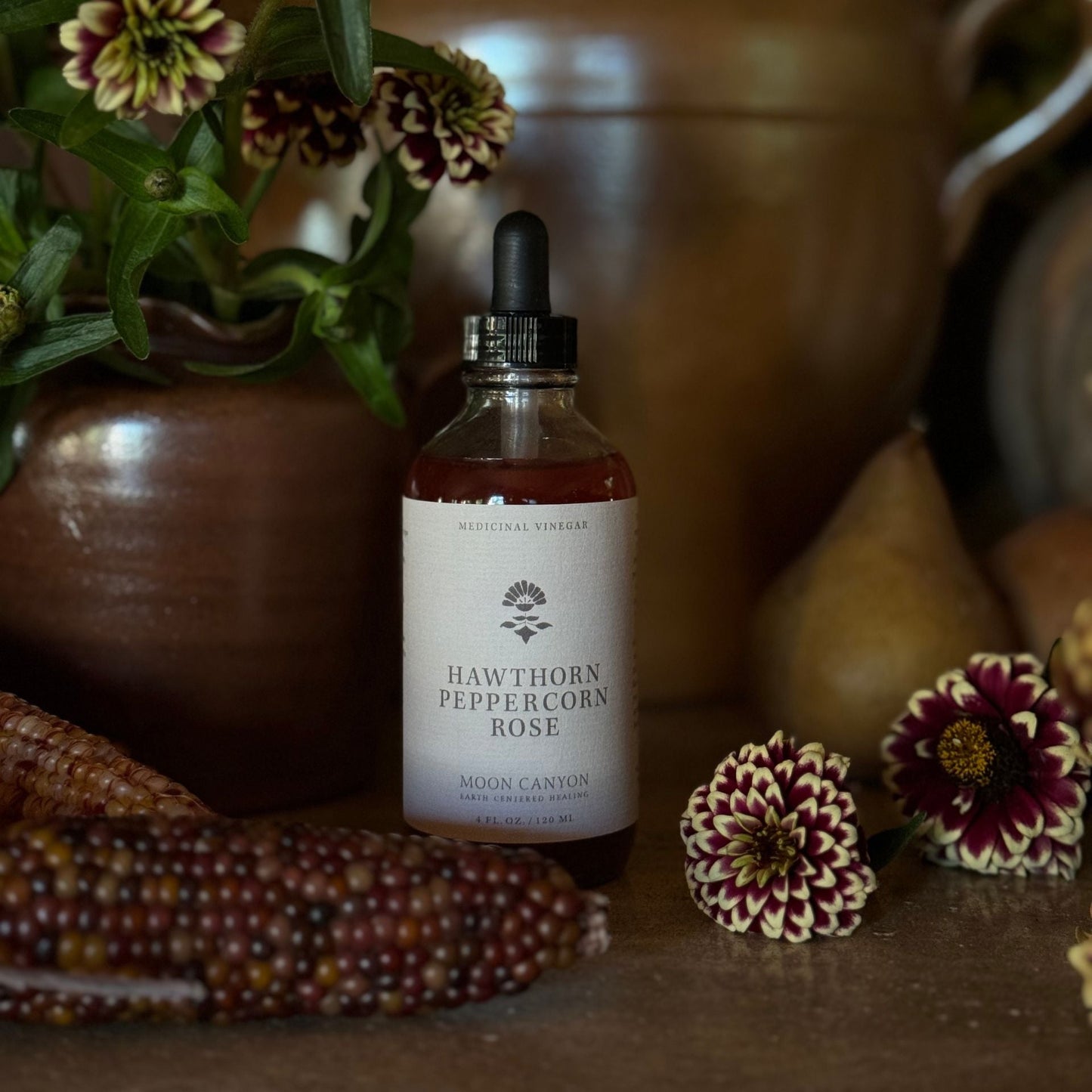 A bottle of Moon Canyon Healing's Hawthorn, Peppercorn & Rose Vinegar with a dropper lid is placed on a wooden surface, surrounded by decorative items such as multicolored corn cobs, flowers, and pears. The background features rustic pots and additional greenery.