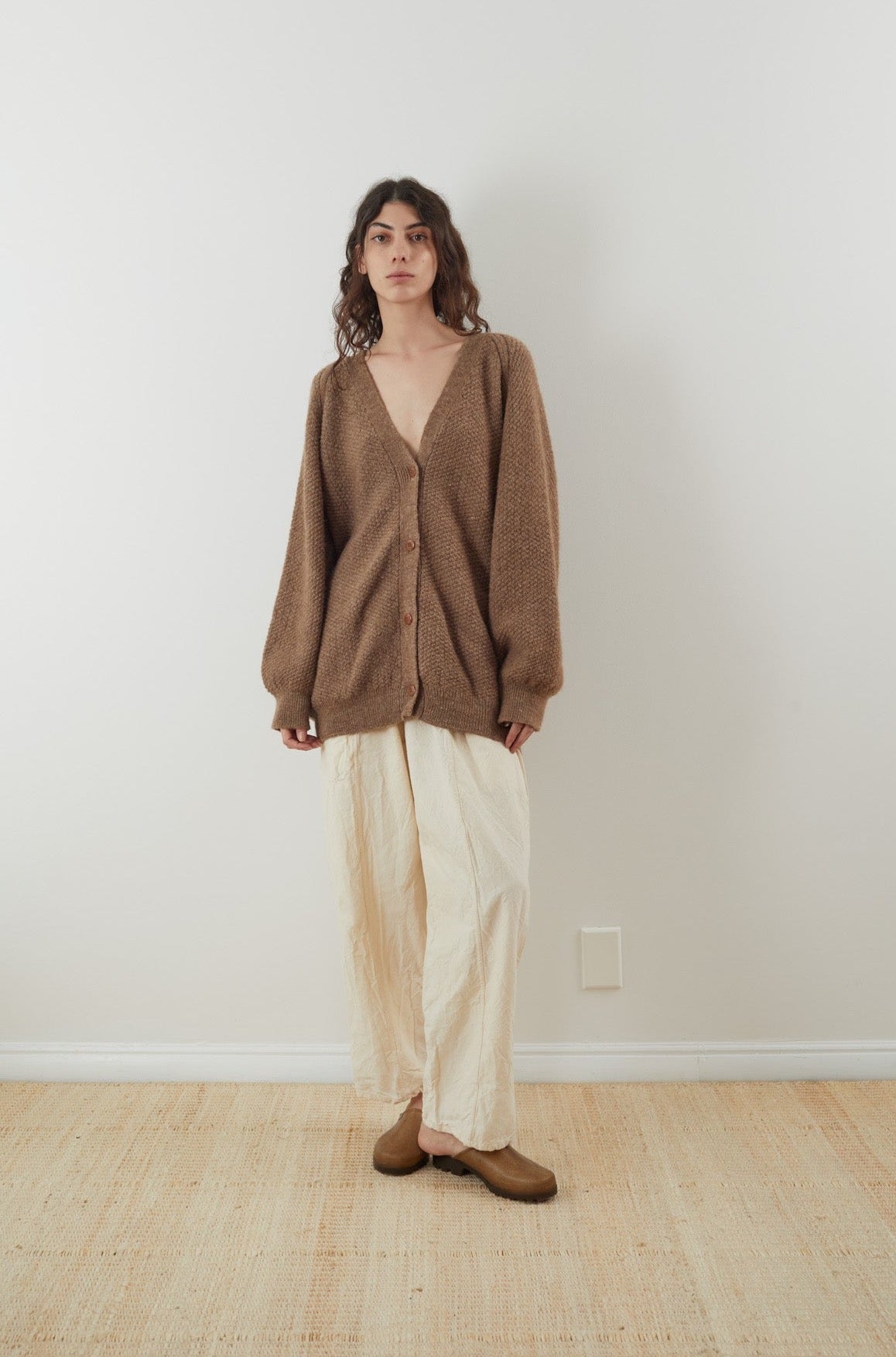 A person with long hair wearing the Wol Hide Moss Cardigan in Earth, cream wide-leg pants, and brown shoes stands against a plain white wall on a woven rug.