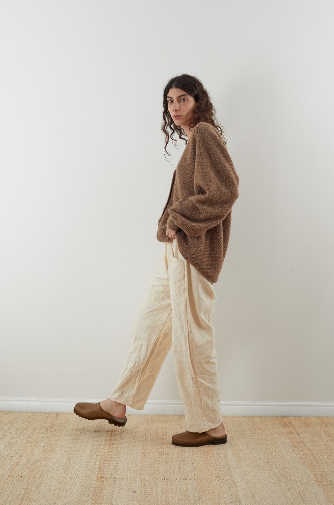 A person with long curly hair stands against a plain backdrop, wearing the oversized Moss Cardigan in Earth from Wol Hide, made from Climate Beneficial wool. Complementing their look are cream pants and brown shoes. The setting is minimalistic, with a light wooden floor enhancing the simplicity.