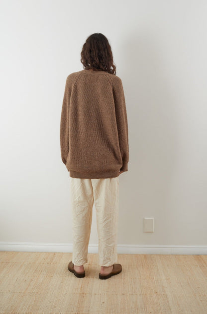 A person with curly hair stands facing a white wall, dressed in Wol Hide's Moss Cardigan in Earth. The oversized fit cardigan, made from climate beneficial wool, is paired with beige pants and brown slippers. The minimalistic room features a light-colored floor and a small electrical outlet on the wall.