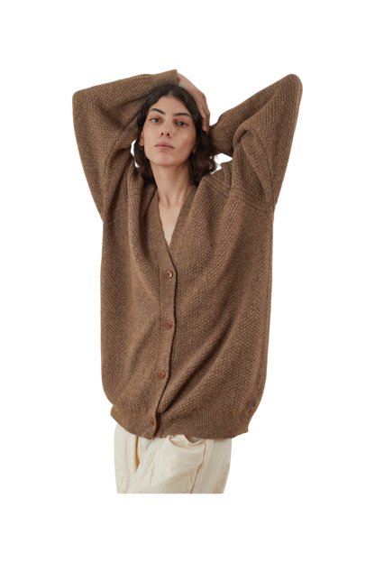 Clad in the Wol Hide Moss Cardigan in Earth, a person with an oversized fit and buttoned front layers it over cream pants, lifting their arms to rest behind their head against a plain white background.