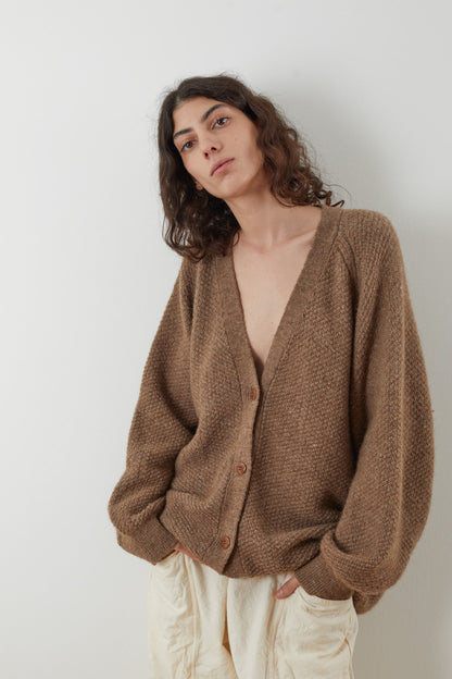 Against a white background, an individual with long, wavy hair models the Moss Cardigan in Earth by Wol Hide. Crafted from Climate Beneficial wool, this oversized brown cardigan features large buttons and is casually layered over cream-colored pants, with the model's hands comfortably resting in their pockets.