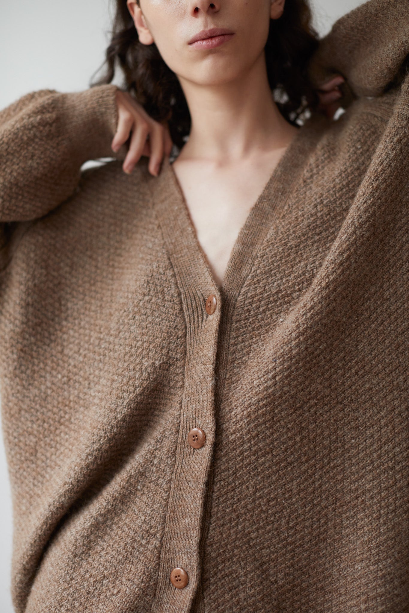 A person wearing the Moss Cardigan in Earth by Wol Hide, an oversized brown knitted cardigan crafted from Climate Beneficial wool, stands with arms slightly raised to display the fabric's texture. The image is cropped at the shoulders and above the knees.