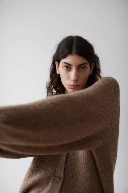 A person with long dark hair is wearing a Moss Cardigan in Earth by Wol Hide, standing in front of a plain white background. One arm is extended toward the camera, partially covering their body.