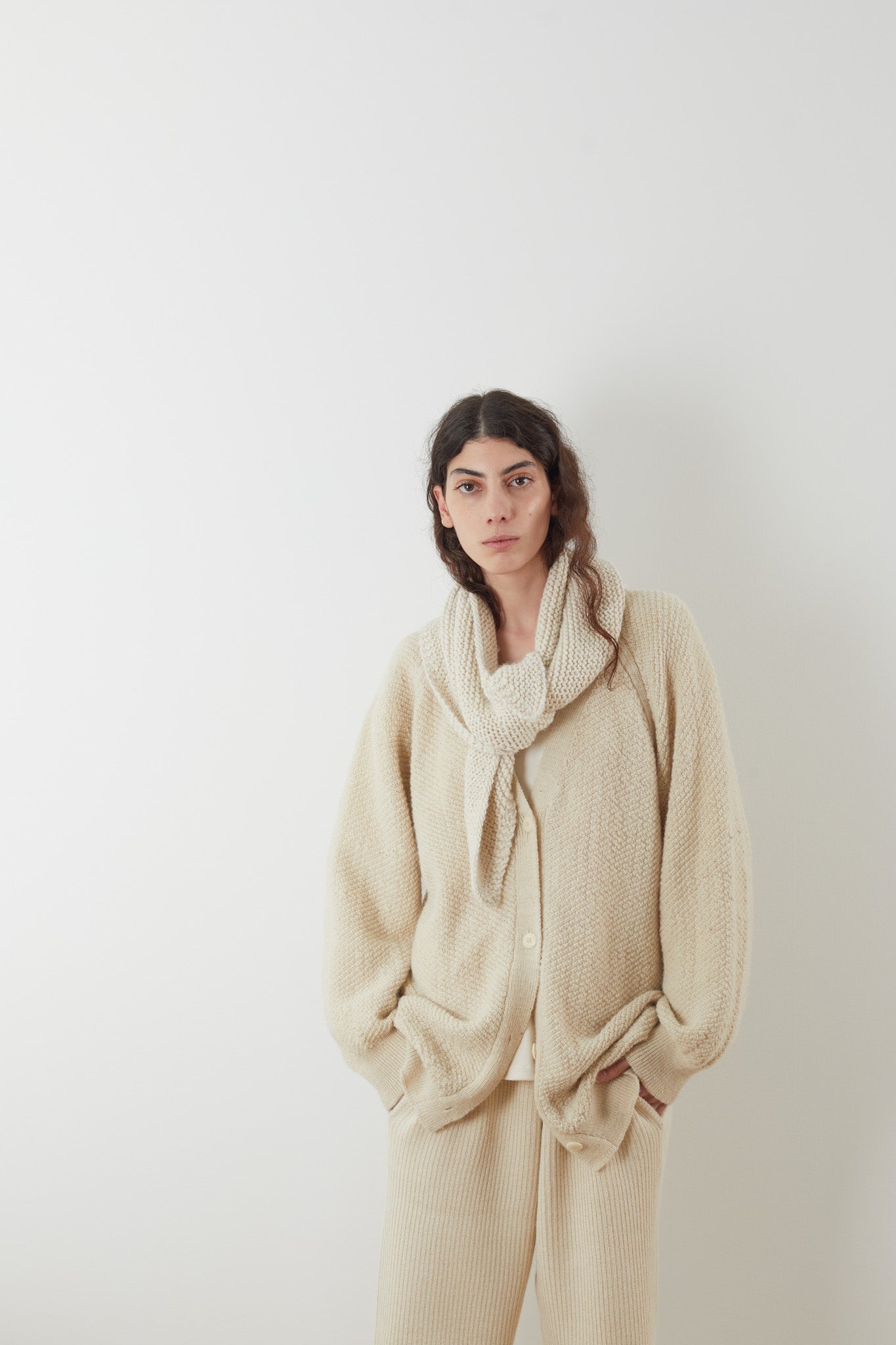 Wrapped in an oversized fit, a person with long hair is wearing the Moss Cardigan in Ecru by Wol Hide, paired with matching pants crafted from Climate Beneficial Alpaca. A cozy scarf drapes around their neck as they tuck their hands into their pockets, standing against a plain white background.