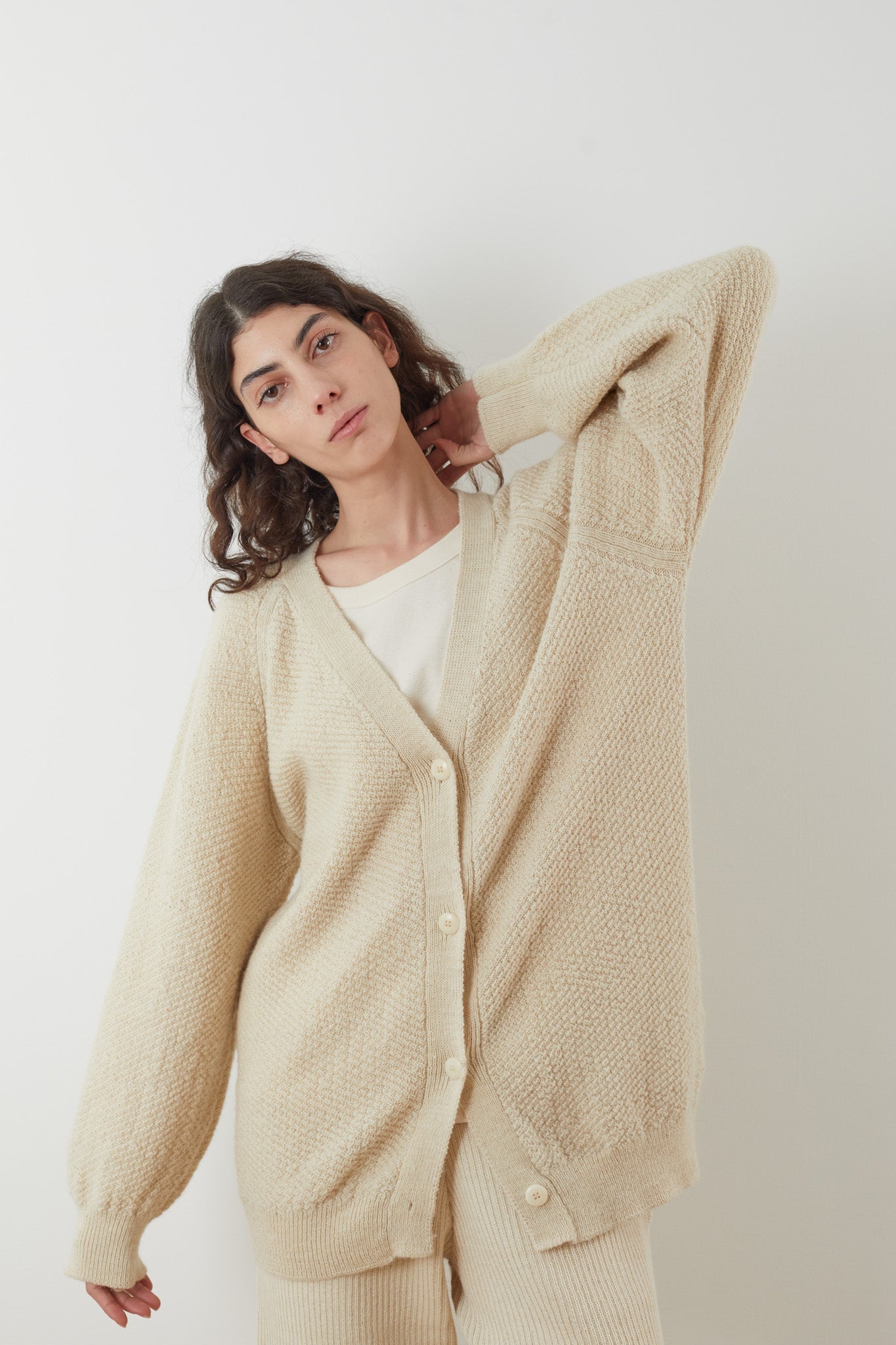 A person with long, curly hair is dressed in the oversized Moss Cardigan in Ecru and matching pants, both made from Climate Beneficial Wool by Wol Hide. They stand against a plain background with one arm raised, touching the back of their head, while looking directly at the camera.