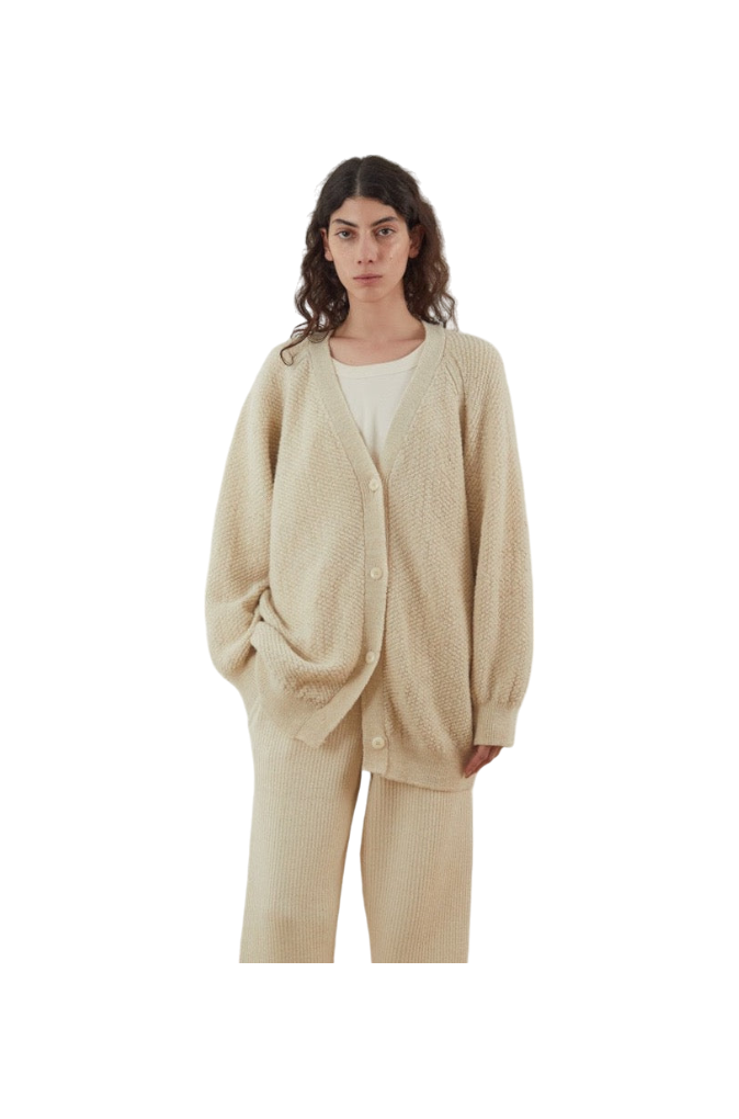 A woman is wearing the Moss Cardigan in Ecru by Wol Hide, an oversized and textured piece made from Climate Beneficial Wool, which she pairs with matching pants and a casual white top. Her long, wavy hair cascades down as she stands against a plain white background.