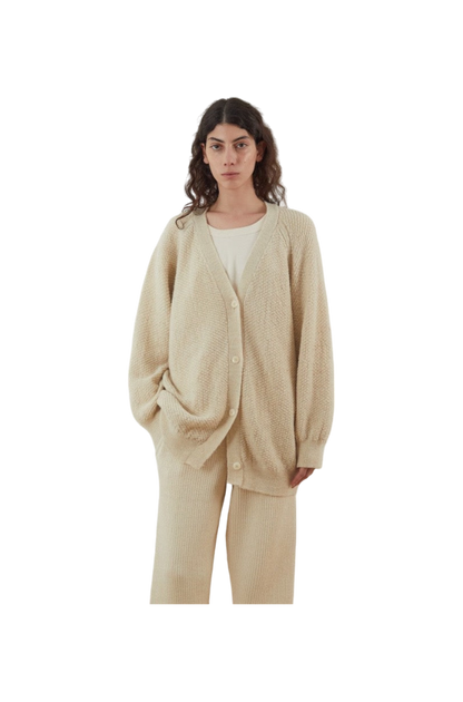 A woman is wearing the Moss Cardigan in Ecru by Wol Hide, an oversized and textured piece made from Climate Beneficial Wool, which she pairs with matching pants and a casual white top. Her long, wavy hair cascades down as she stands against a plain white background.