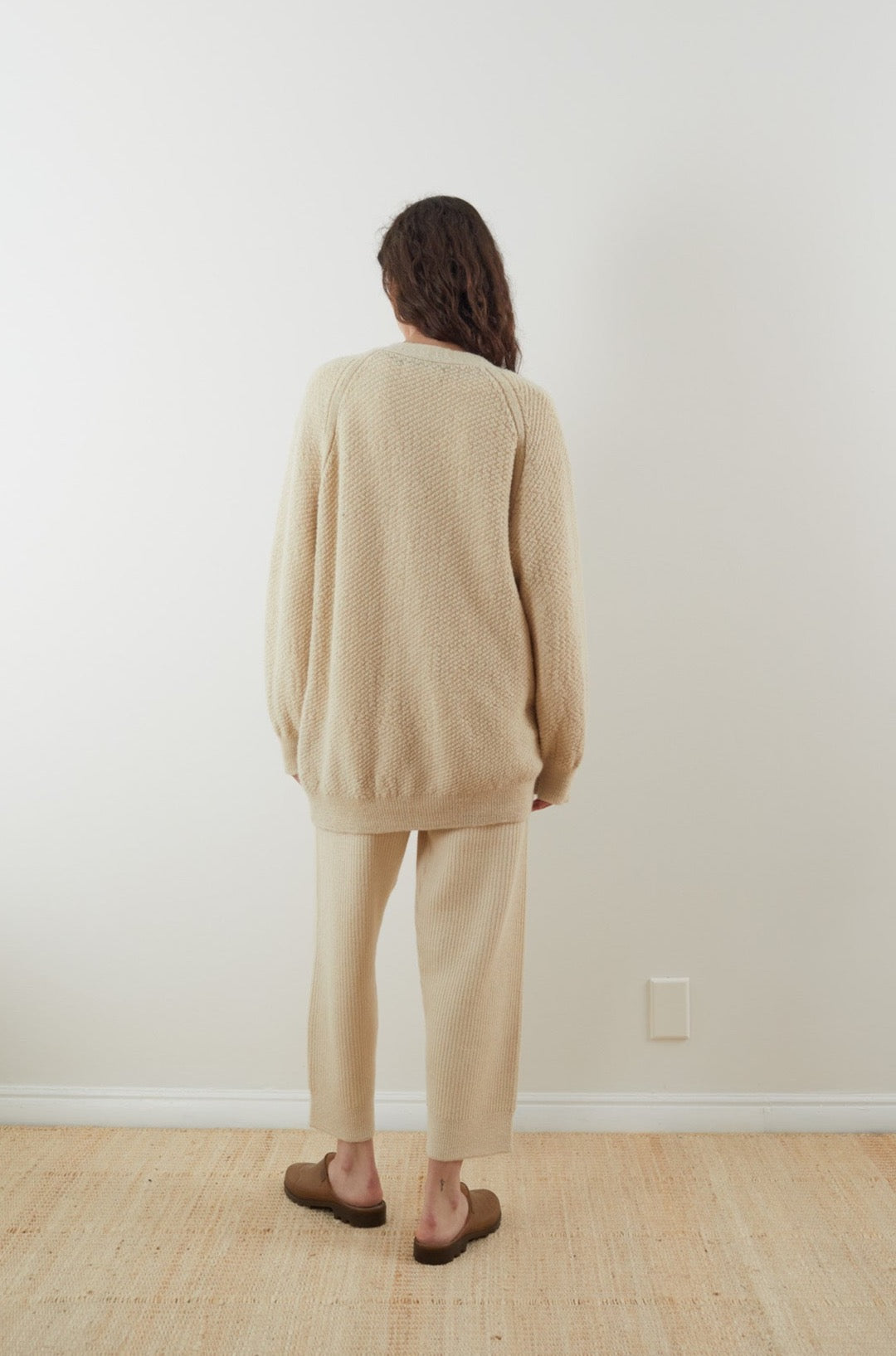 A person with long hair stands facing a plain white wall, wearing the cozy oversized fit Moss Cardigan in Ecru by Wol Hide, paired with matching beige pants and brown slip-on shoes. They stand on a light-colored rug in a minimalist setting.