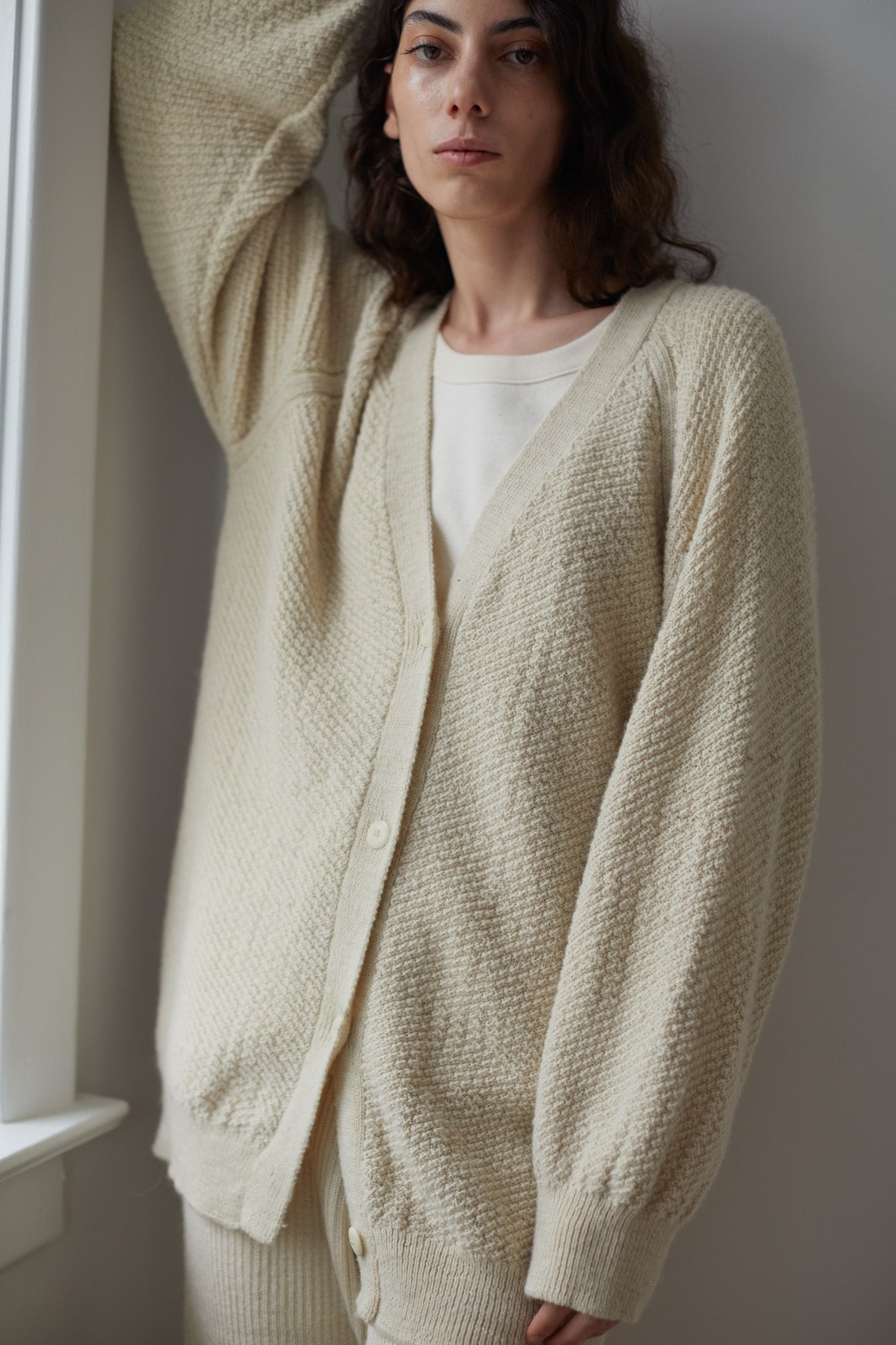 A person with long hair stands leaning against a wall by a window, wearing Wol Hide's oversized Moss Cardigan in Ecru over a matching top. Made from Climate Beneficial Wool, the cardigan features a textured, knitted design with buttons. The setting appears to be indoors.