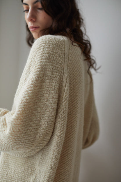 A person with long, dark hair is wearing the Moss Cardigan in Ecru, an oversized fit, textured light beige knit sweater from Wol Hide, crafted from Climate Beneficial Wool. The detailed fabric of the cardigan is the focal point as the person gazes downward with a calm expression against a neutral background.