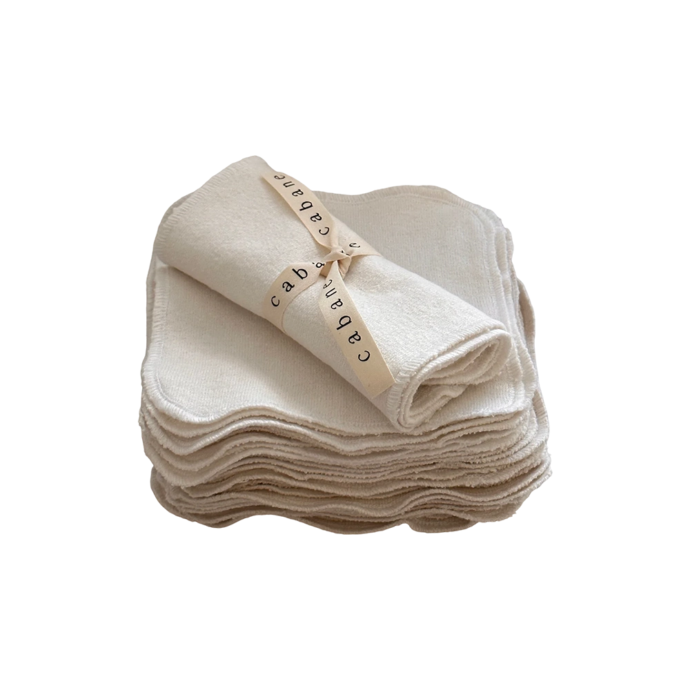 A stack of neatly folded beige cloth washcloths, crafted from undyed organic cotton, with one washcloth rolled up and secured with a cream-colored ribbon labeled "cabane childrenswear." The stack is arranged in an orderly manner on a white surface, embodying the promise of a gentle washing experience. This is the Organic Washcloth Set of 4 from cabane childrenswear.