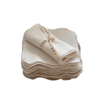 A stack of neatly folded beige cloth washcloths, crafted from undyed organic cotton, with one washcloth rolled up and secured with a cream-colored ribbon labeled "cabane childrenswear." The stack is arranged in an orderly manner on a white surface, embodying the promise of a gentle washing experience. This is the Organic Washcloth Set of 4 from cabane childrenswear.