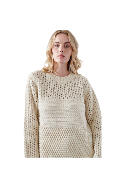 A person is wearing the Wol Hide Mixed Stitch Pullover in Ecru, an oversized knit sweater crafted from alpaca wool. They stand against a solid black background with shoulder-length blonde hair and look directly at the camera.