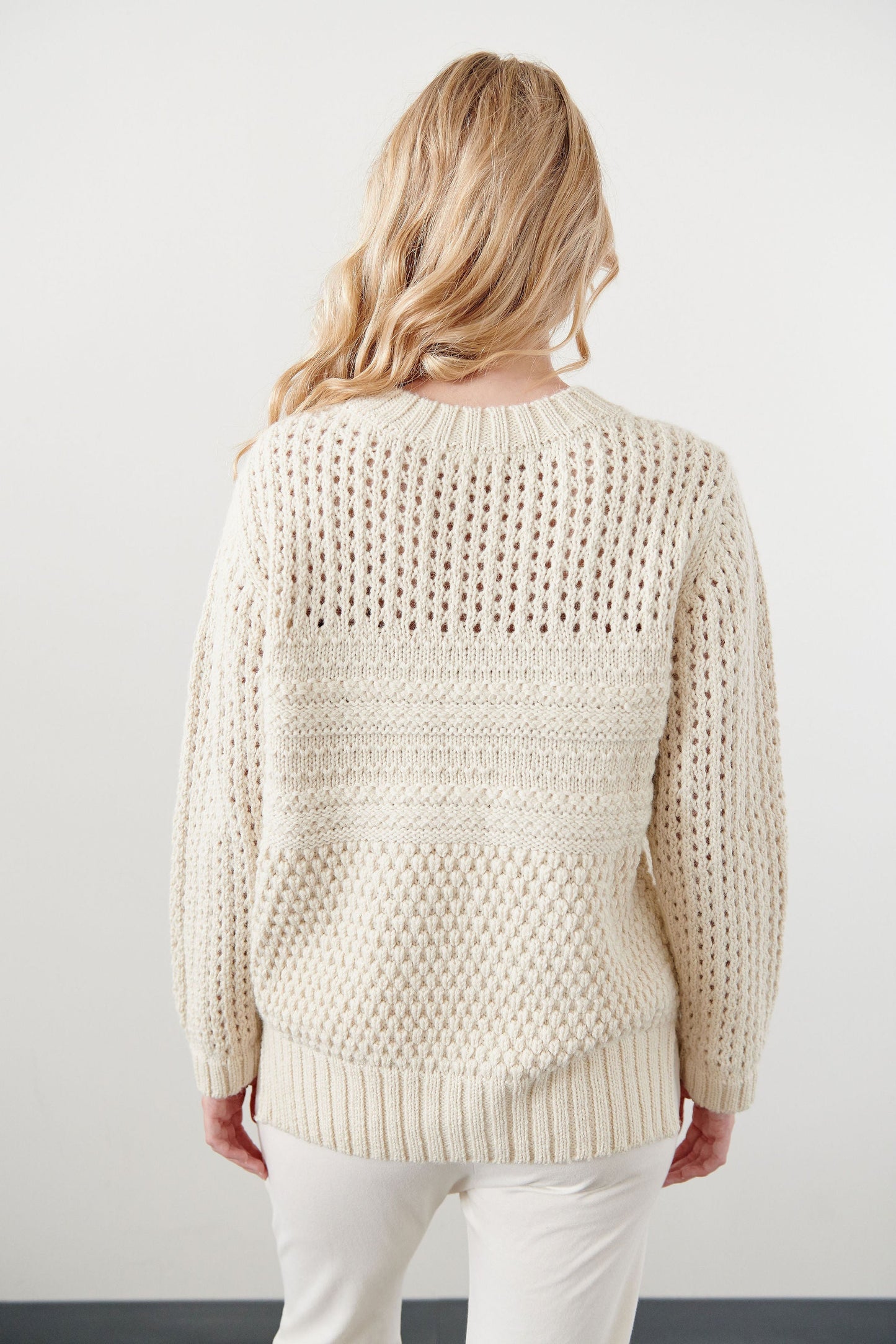 A person with long, wavy blonde hair stands with their back to the camera, wearing the Wol Hide Mixed Stitch Pullover in Ecru over white pants. The backdrop is a plain off-white wall.