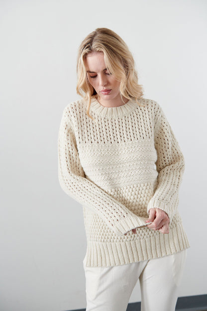 A person with shoulder-length blonde hair stands against a plain, light-colored wall, wearing the Mixed Stitch Pullover in Ecru by Wol Hide and white pants, looking down with a relaxed expression.