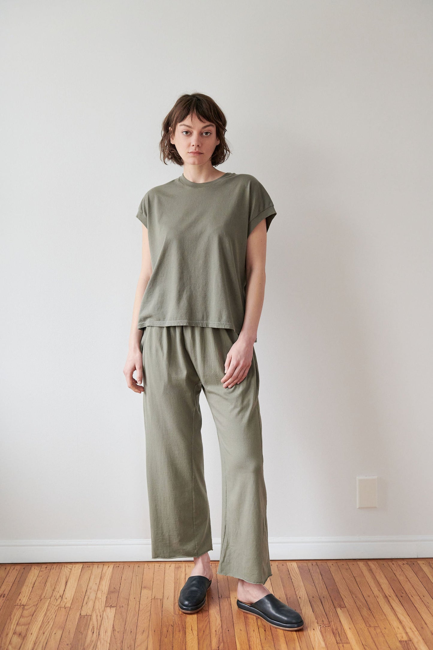 A person with short brown hair stands in a minimalistic room with a wooden floor and white walls, wearing Wol Hide's Organic Cotton Jersey Box Tee in Fir with matching pants and black slip-on shoes. Made in the USA, their look is complemented by a neutral expression.