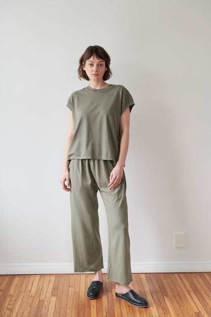 A person with short brown hair stands in a minimalistic room with a wooden floor and white walls, wearing Wol Hide's Organic Cotton Jersey Box Tee in Fir with matching pants and black slip-on shoes. Made in the USA, their look is complemented by a neutral expression.