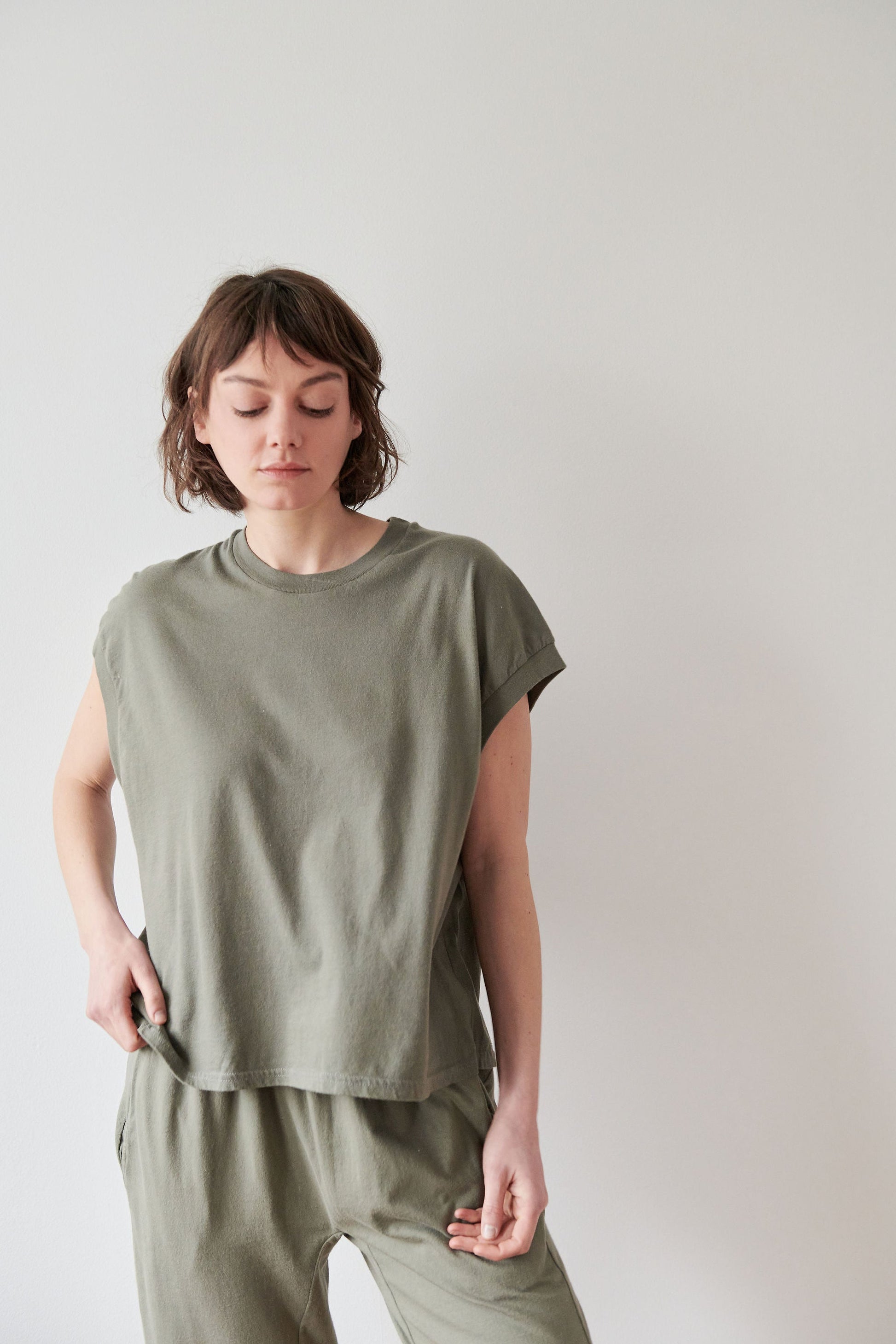 A short brown-haired individual stands against a white background, wearing Wol Hide's Organic Cotton Jersey Box Tee in Fir and matching olive-green pants. Made from organic cotton and crafted in the USA, they look down with a hand in their pocket.