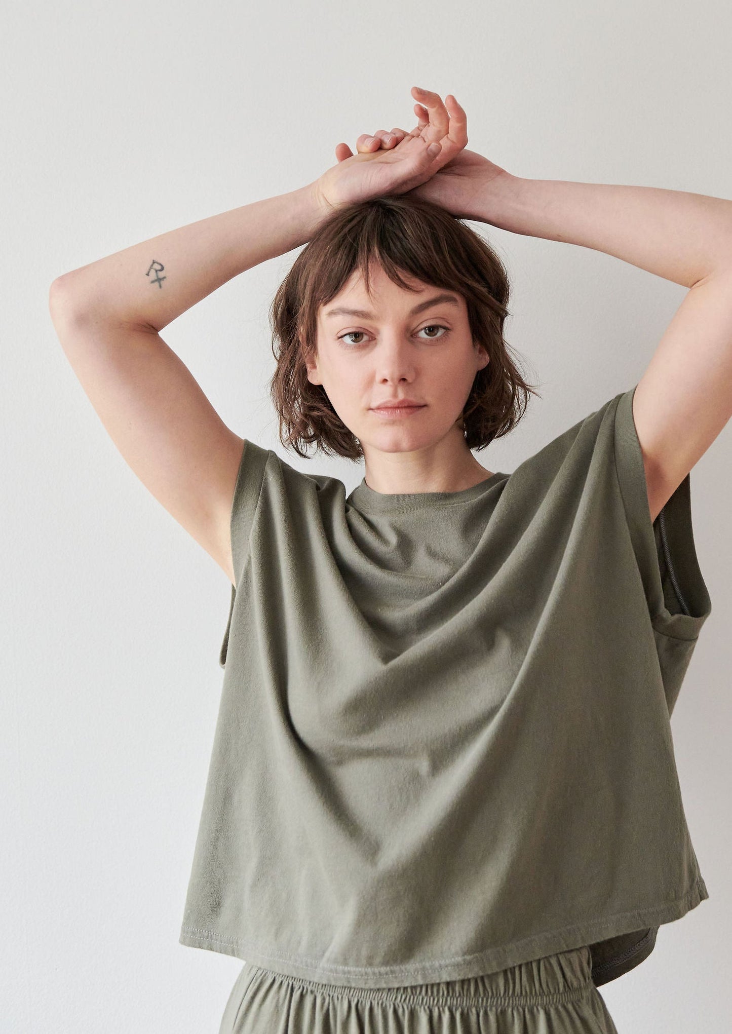 A person with short brown hair and a neutral expression stands against a plain background, wearing the Wol Hide Cotton Jersey Box Tee in Fir. They have their arms raised above their head, revealing a tattoo on their right forearm.