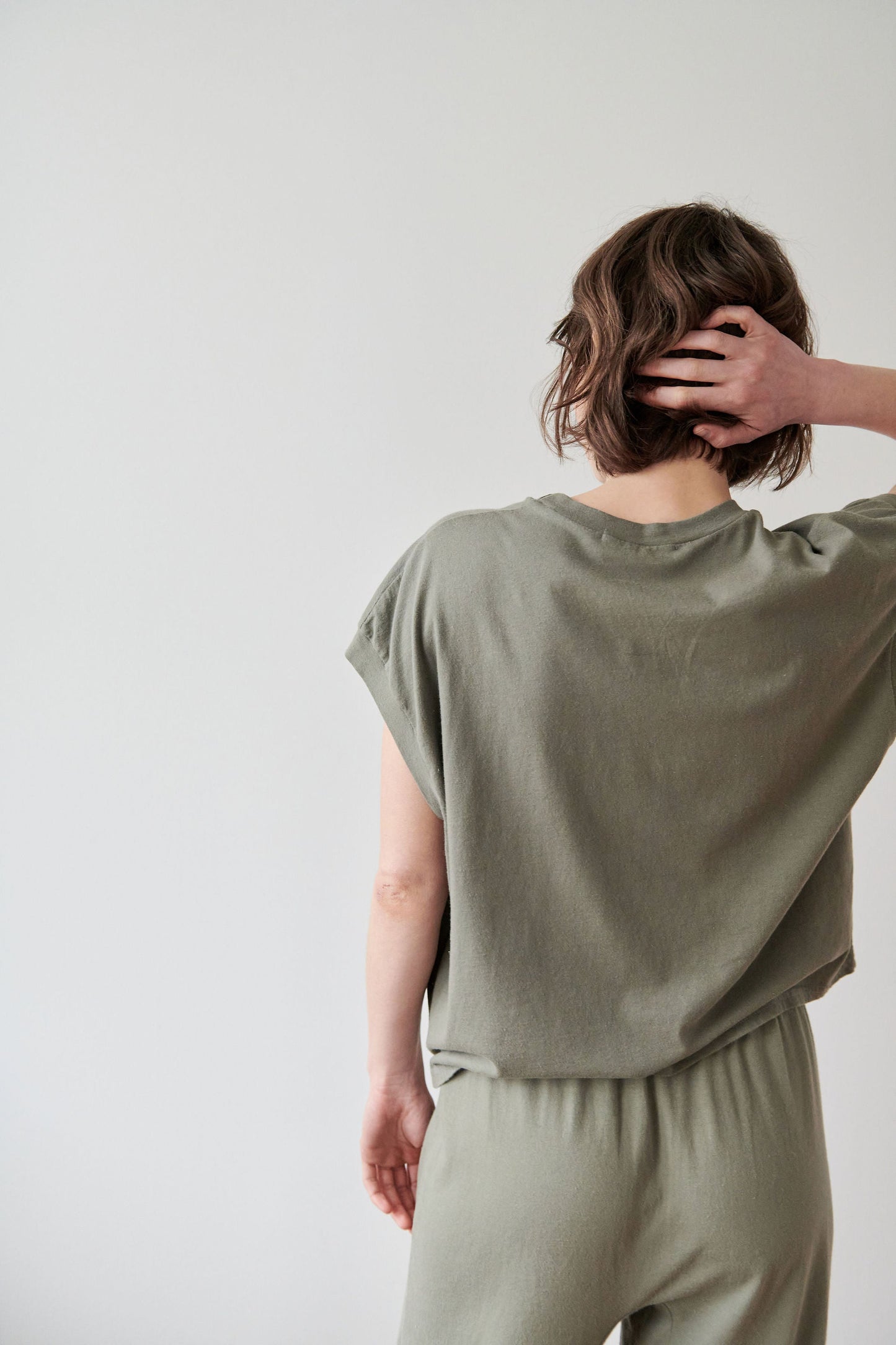 A person with short hair faces away, wearing the Wol Hide Organic Cotton Jersey Box Tee in Fir and matching pants, their right hand in their hair against a plain white background.