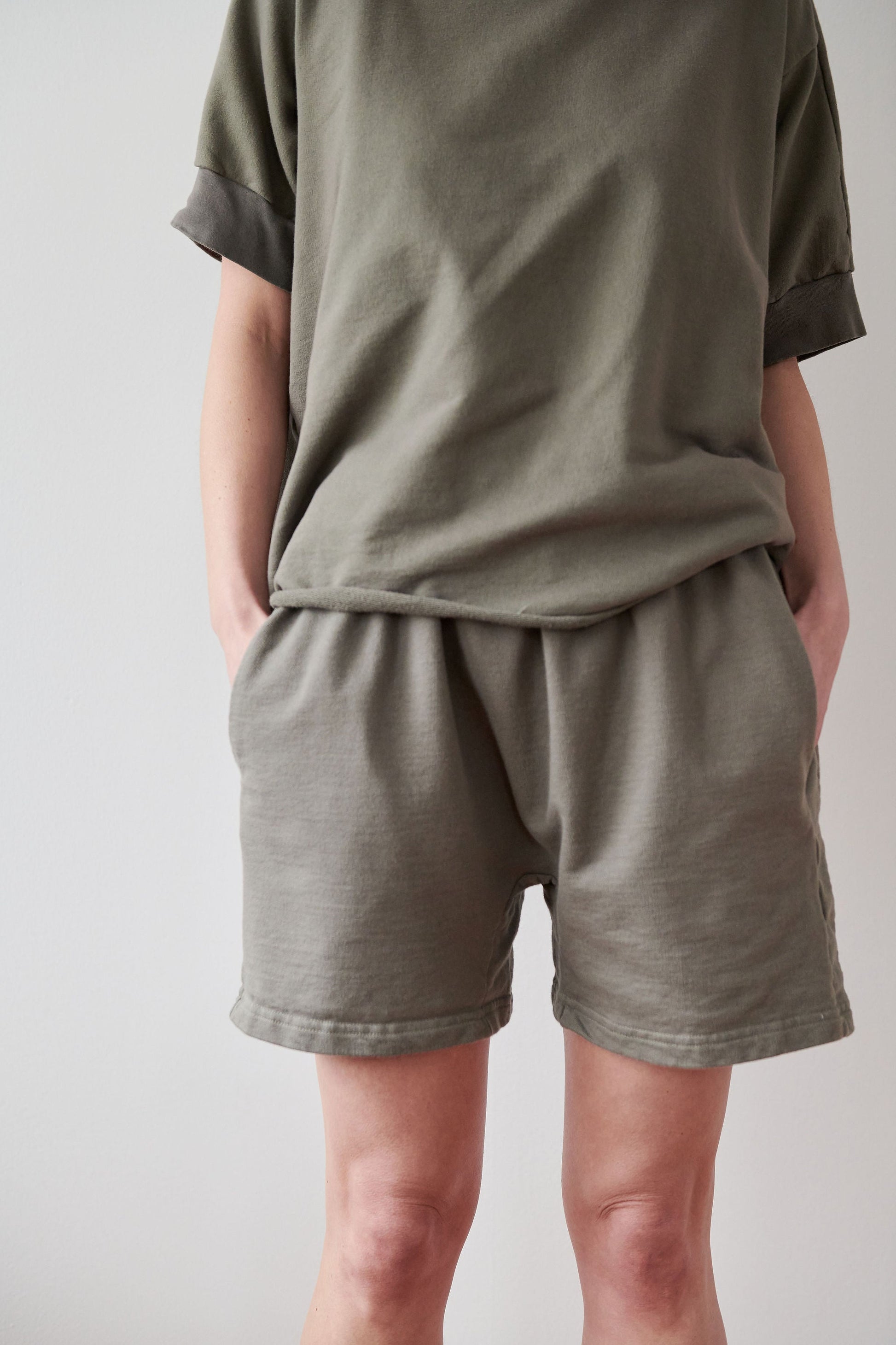 A person wearing an oversized olive green t-shirt and matching Wol Hide's Cotton Easy Shorts in Fir, featuring a high-rise elastic waist, stands with their hands in their pockets against a plain light-colored background. The person's face is not visible in the image, and the outfit is proudly made in the USA.