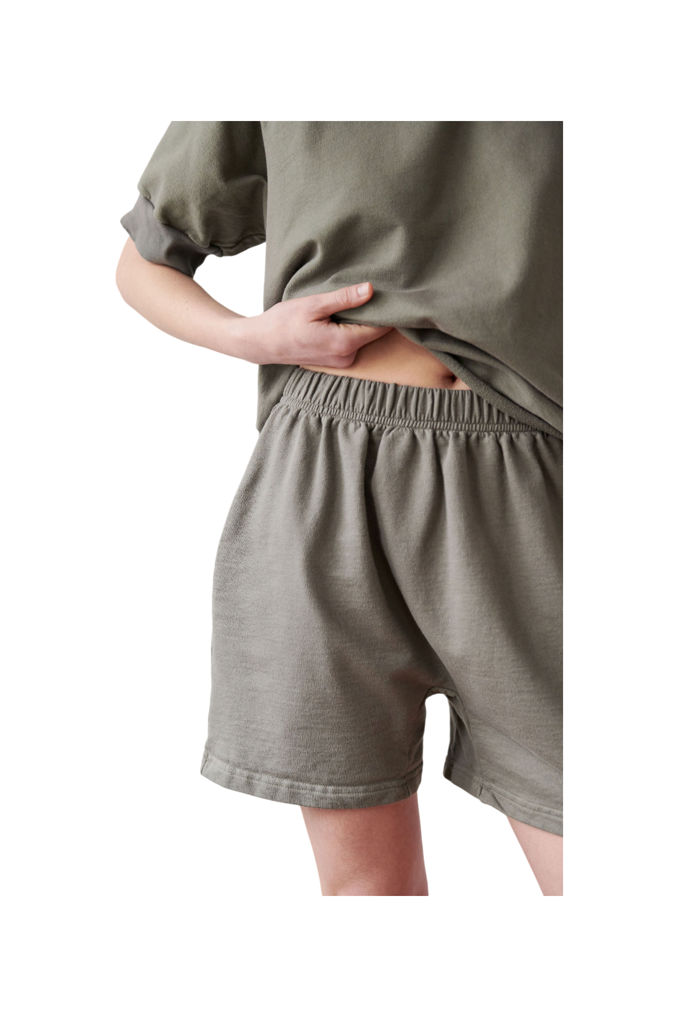 A person wearing loose-fitting, beige Cotton Easy Shorts in Fir from Wol Hide is seen from mid-thigh to mid-torso. The individual slightly lifts their T-shirt to reveal the high-rise elastic waistband of the shorts. The background is neutral. 