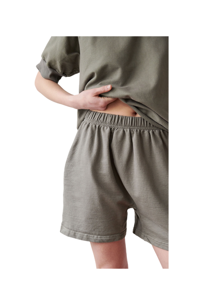 A person wearing loose-fitting, beige Cotton Easy Shorts in Fir from Wol Hide is seen from mid-thigh to mid-torso. The individual slightly lifts their T-shirt to reveal the high-rise elastic waistband of the shorts. The background is neutral. 