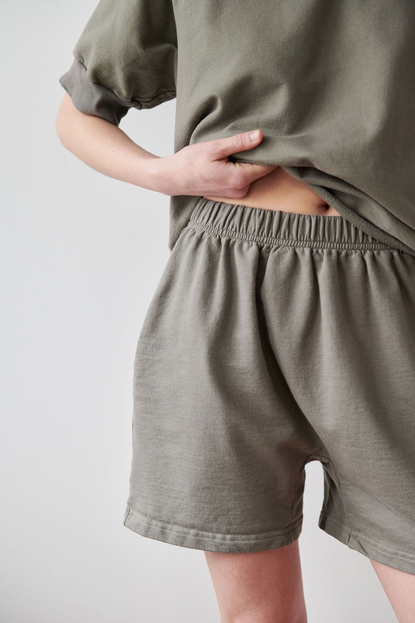 A person wearing a greenish-grey outfit featuring a short-sleeved top and Wol Hide's Cotton Easy Shorts in Fir. The person is partially lifting the top to reveal the high-rise elastic waist of the shorts. The background is a light, neutral color, complementing the casual elegance of their Made in USA ensemble.