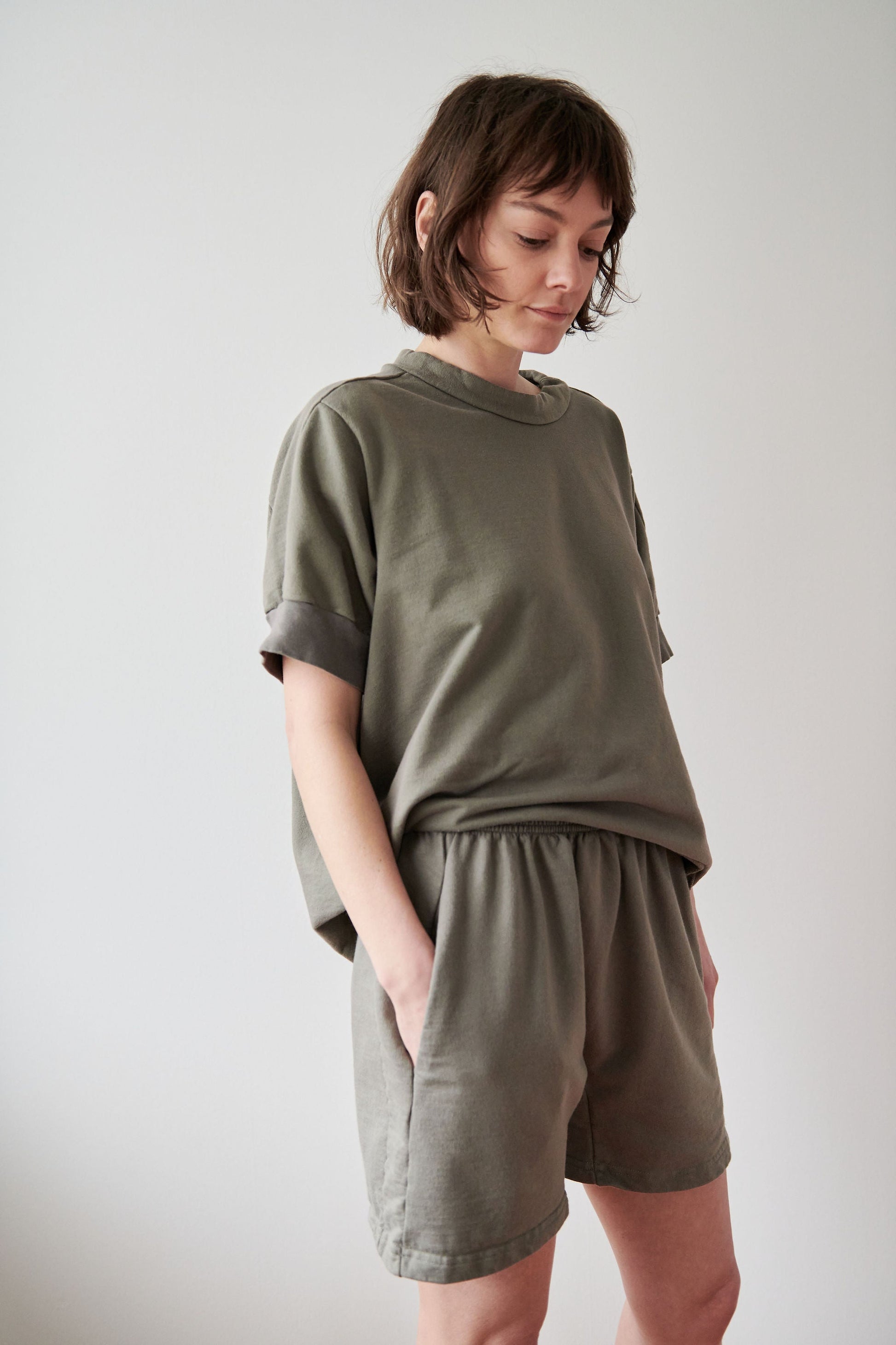 A person with short, tousled hair is looking down while standing against a plain background. They are wearing a loose-fitting, olive green short-sleeved top and Wol Hide's Cotton Easy Shorts in Fir, which feature a high-rise elastic waist and hands in their pockets. The overall mood appears pensive or introspective.