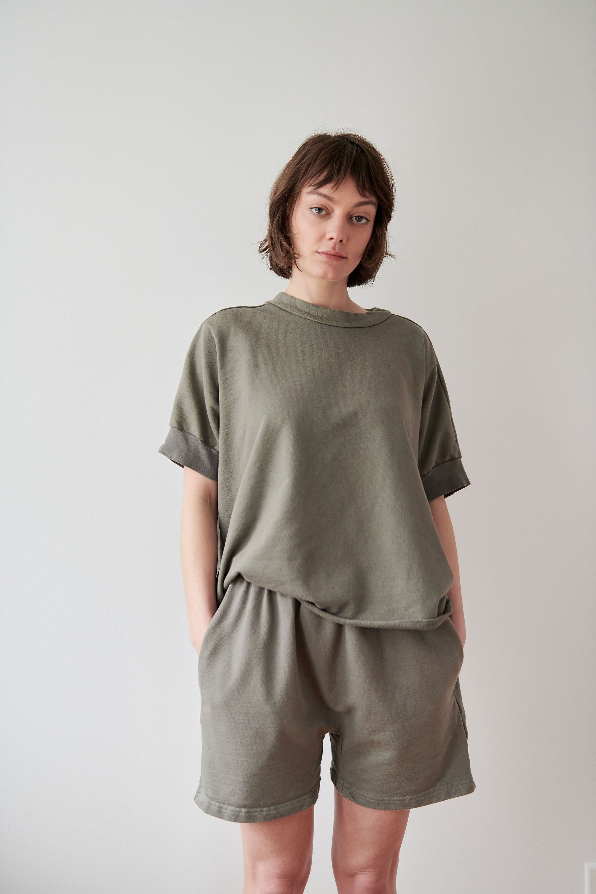 A person with short brown hair stands against a plain light-colored background. They are wearing a loose, olive-green t-shirt and Wol Hide's high-rise elastic waist, Cotton Easy Shorts in Fir with their hands in the pockets. Made in USA, they have a neutral expression and are looking slightly downward.